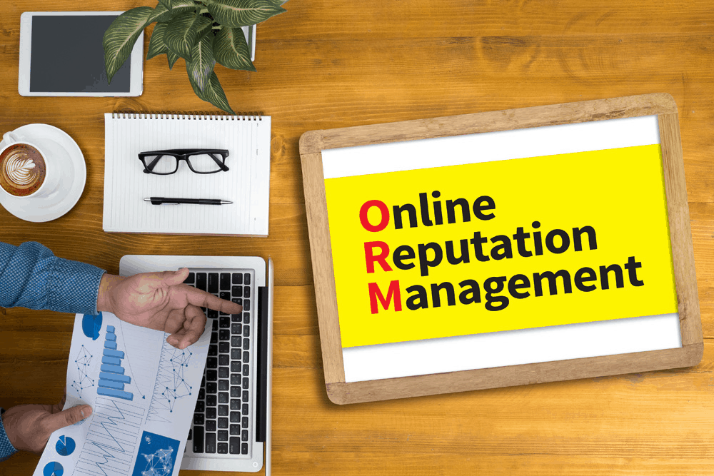 Yellow sheet on a wooden table saying 'Online Reputation Management' listed below one another with the first letter of each word in red. Glasses and a pen on a white notepad with a coffee and someone's hands visible on a latop keyboard