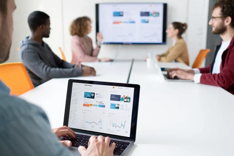 Marketing specialist reviewing A/B testing results on laptop while team analyzes campaign data on wall display